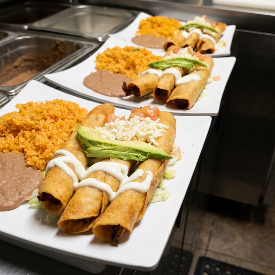 Plates of golden taquitos topped with crema and avocado, served with Mexican rice and refried beans at a restaurant counter.