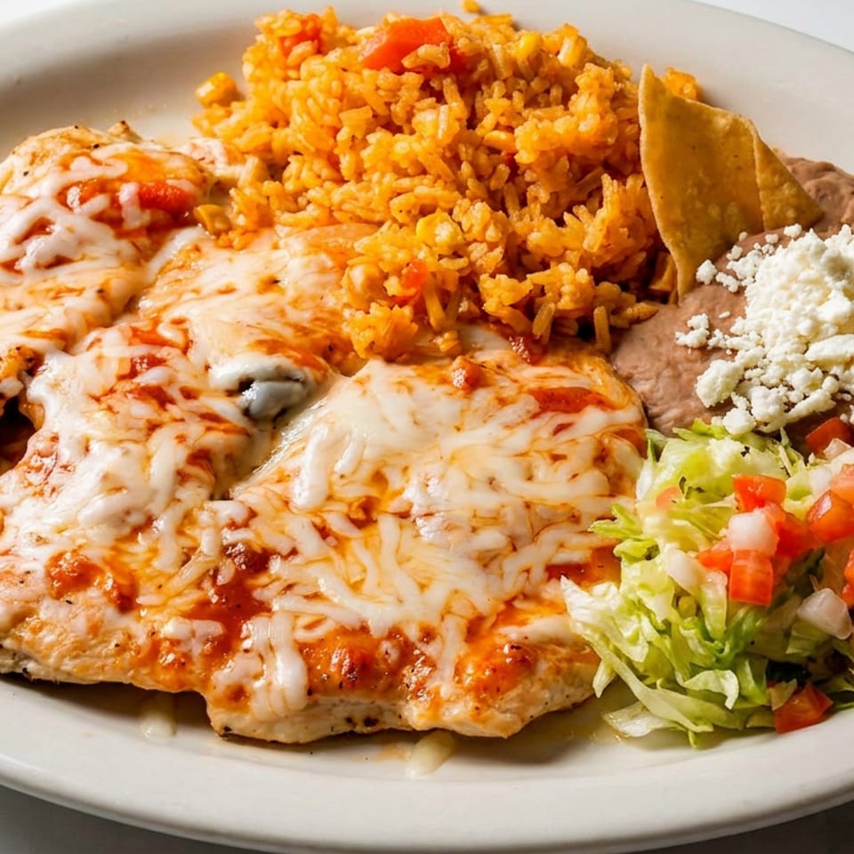 Plate of cheese-topped grilled chicken with Mexican rice, refried beans, tortilla chips, and lettuce salad.